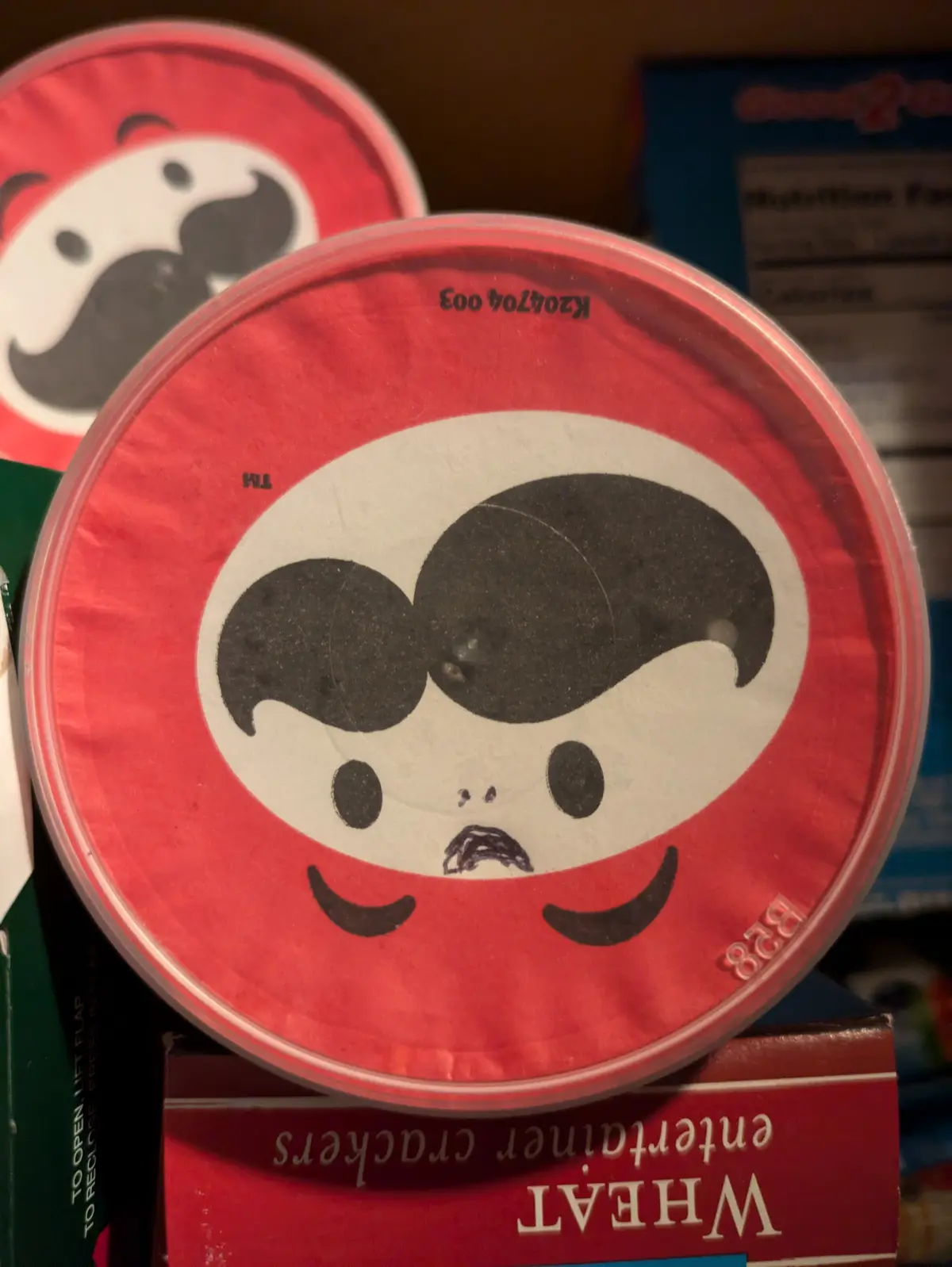 A close-up photo shows a red circular container lid with a cartoon face featuring a black mustache, black eyes, and a…