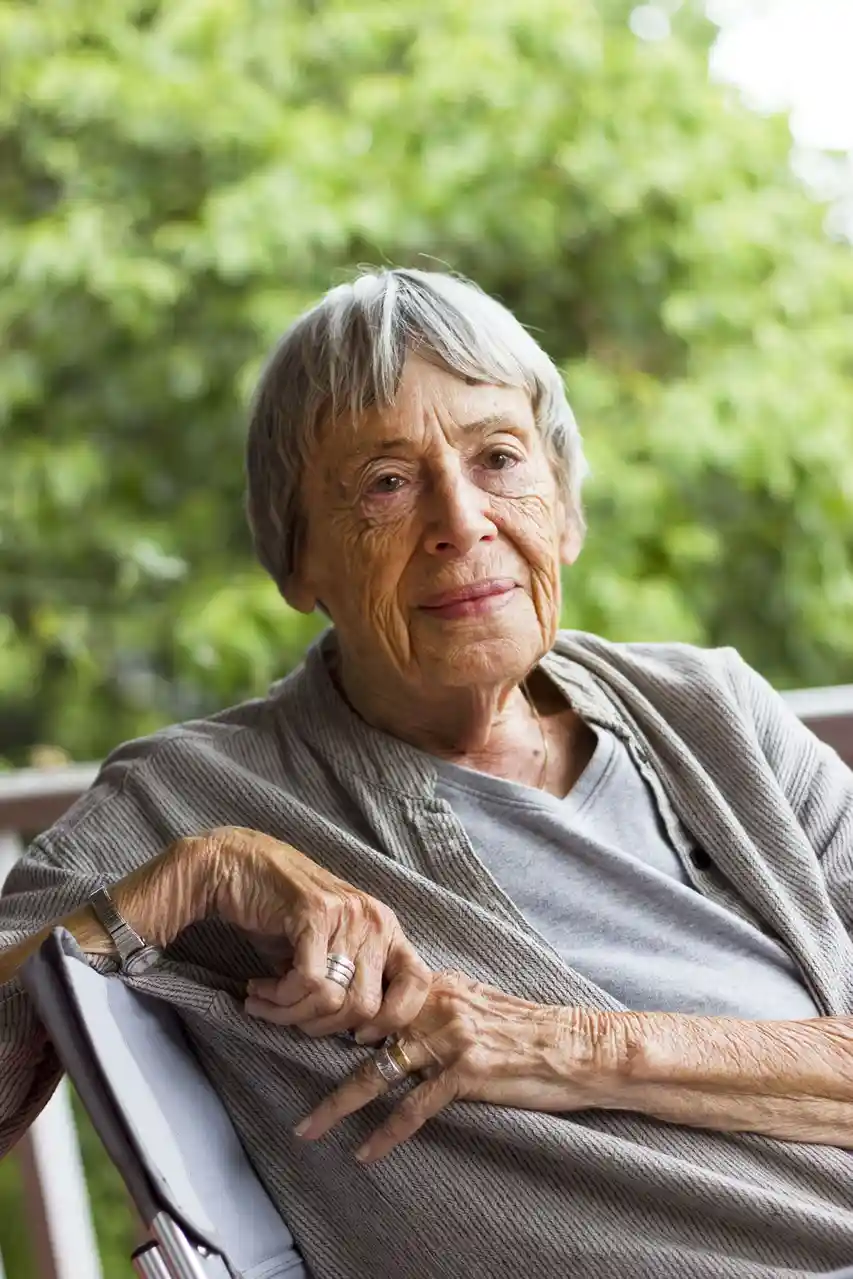 An elderly woman with gray hair sits calmly in a chair outdoors, wearing a gray cardigan over a t-shirt, with a gentle…