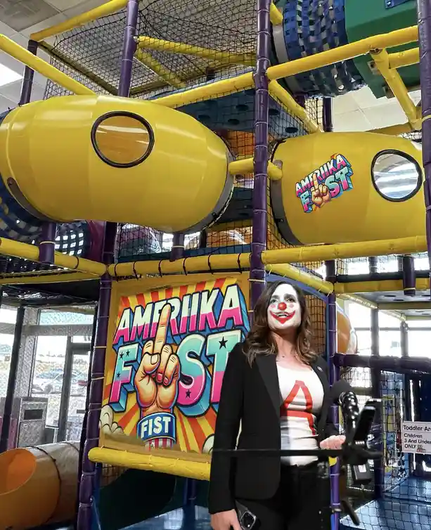 A woman with clown makeup stands at a podium in front of a colorful indoor playground structure. The playground features…