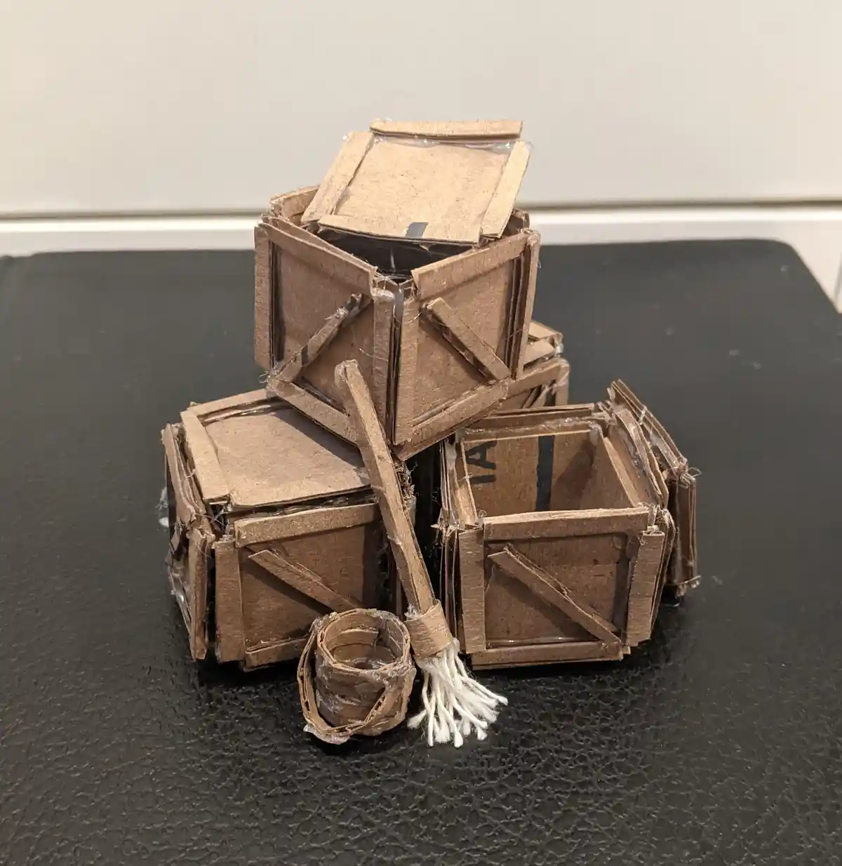 A handmade miniature model of several cardboard crates, some stacked and one tipped over, with a tiny broom and bucket…