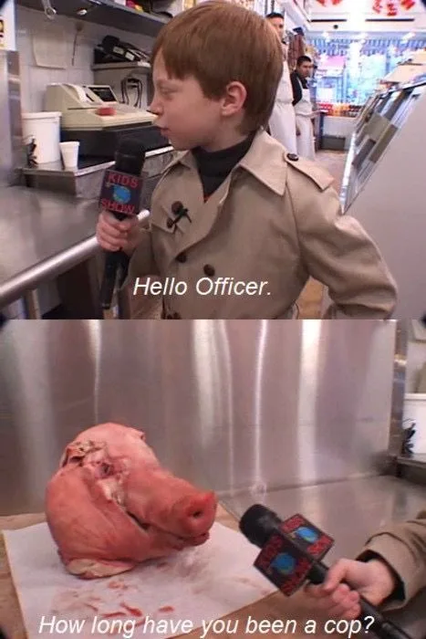 A young boy in a tan trench coat holds a microphone labeled 'KIDS SHOW' and addresses a pig's head on a butcher block…