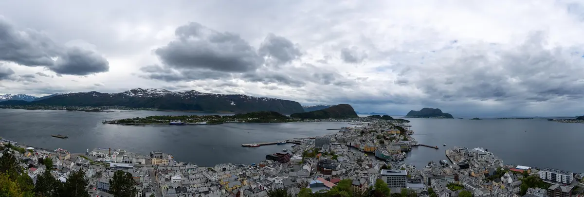 A panoramic view of a coastal town with colorful buildings nestled along a fjord, surrounded by mountains with snow-capped…