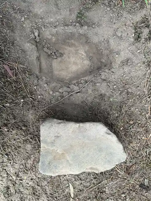 An overhead shot shows a roughly rectangular hole dug into dirt, with a flat, gray rock resting just outside the hole's…