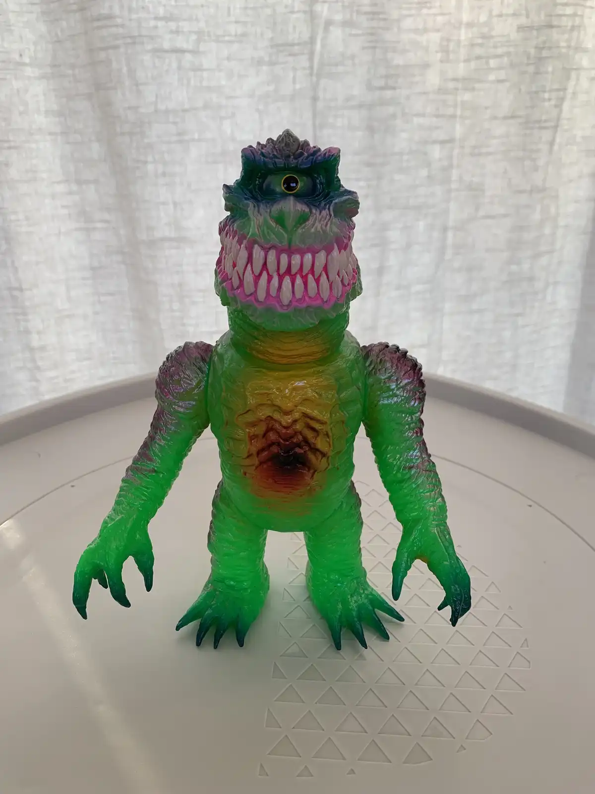 A green, multi-colored plastic monster toy with sharp teeth, clawed hands and feet, and a single glowing eye stands on a…