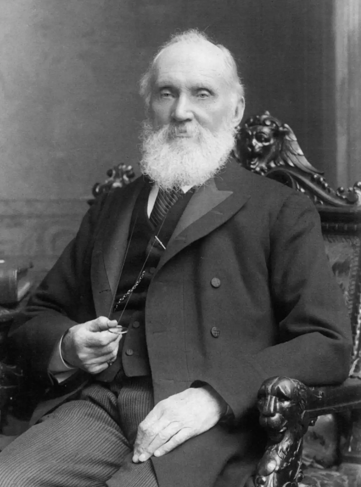 A black-and-white portrait of an elderly man with a long white beard, wearing a formal suit and holding a monocle, seated in…