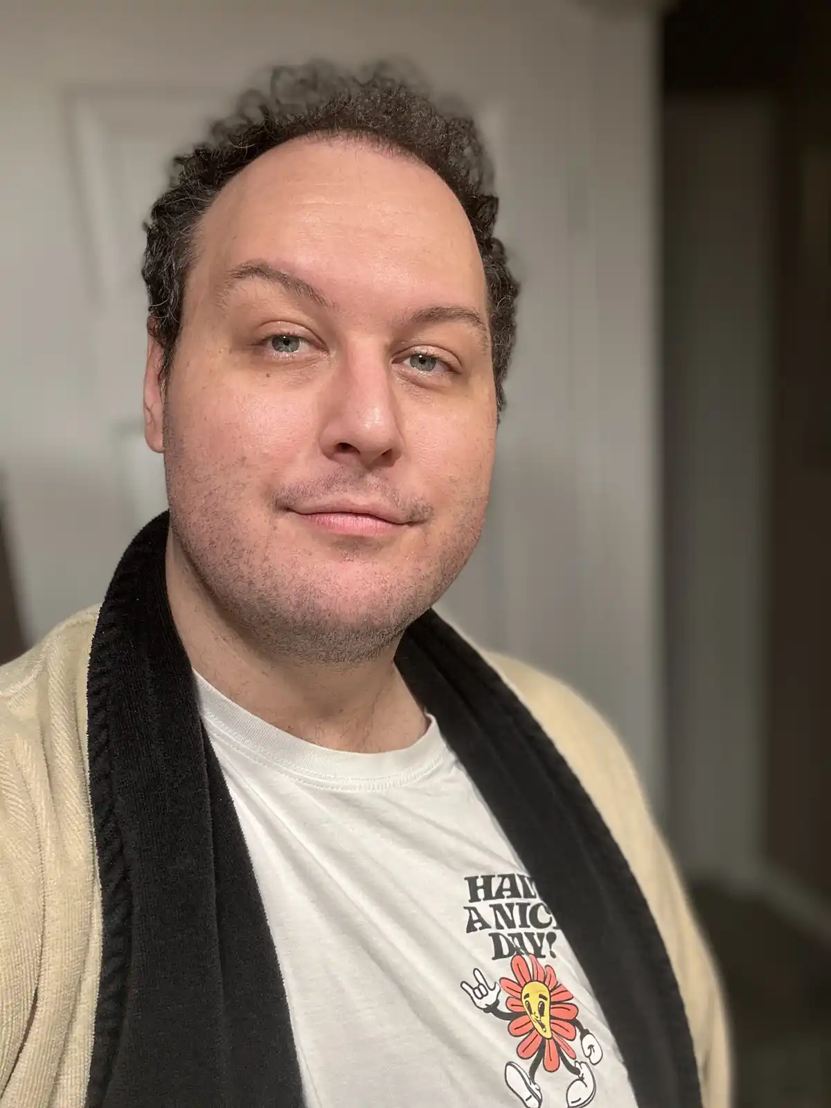 A man with curly dark hair and light eyes looks at the camera with a subtle, slightly smug smile. He wears a white t-shirt…