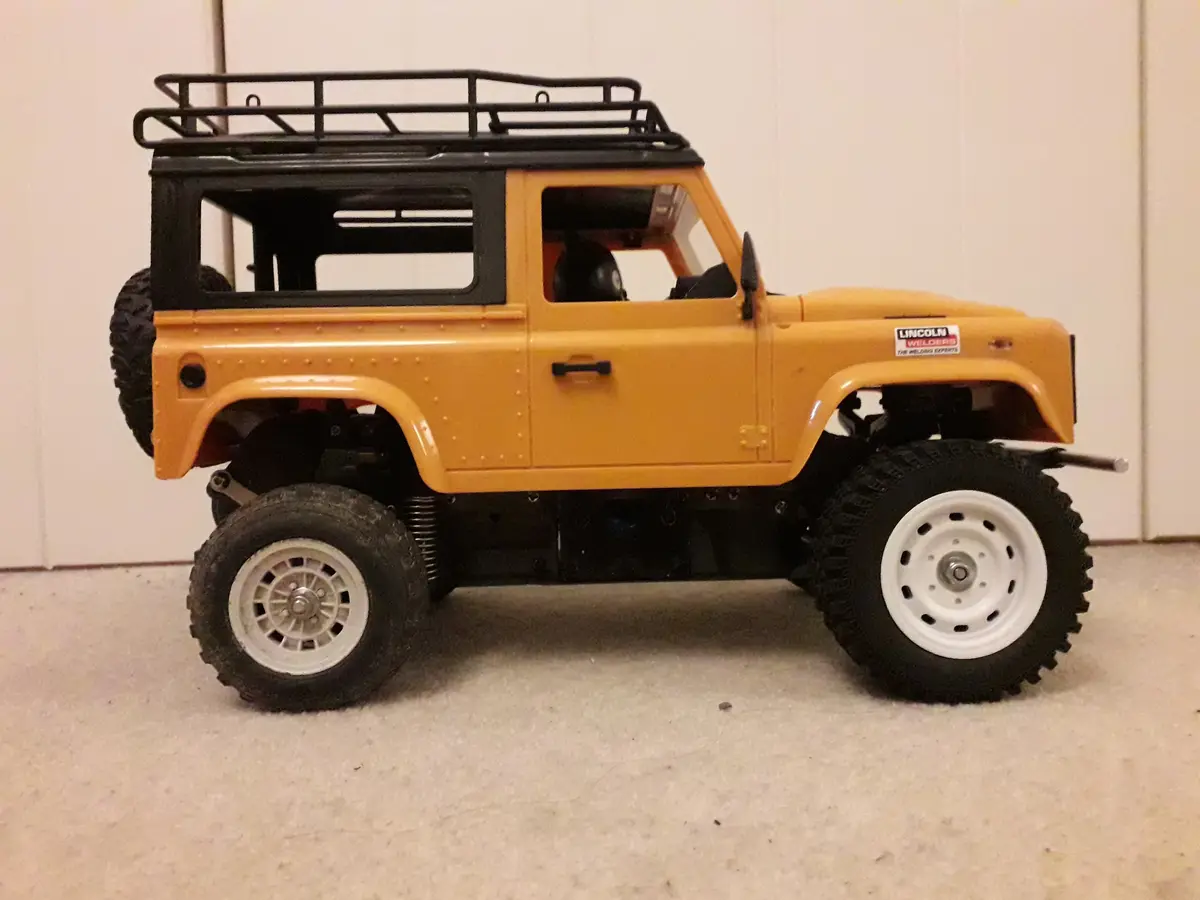 A yellow remote-controlled model of a Land Rover Defender with a black roof rack and white-rimmed tires, shown in profile…