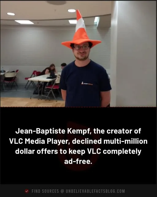 A man wearing an orange and white traffic cone hat stands in a room with tables and chairs, smiling at the camera. The text…