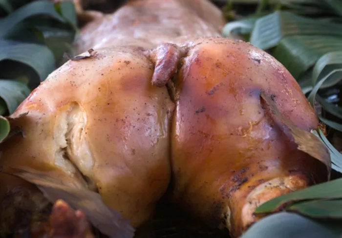 a close-up photo of a roasted pig, its skin glistening with a golden-brown crust, resting on green banana leaves. The pig's…