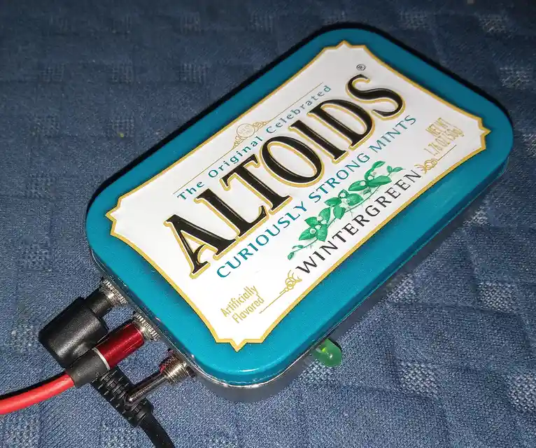 A teal Altoids wintergreen mint tin is repurposed as a portable electronic device, with red and black wires and connectors…