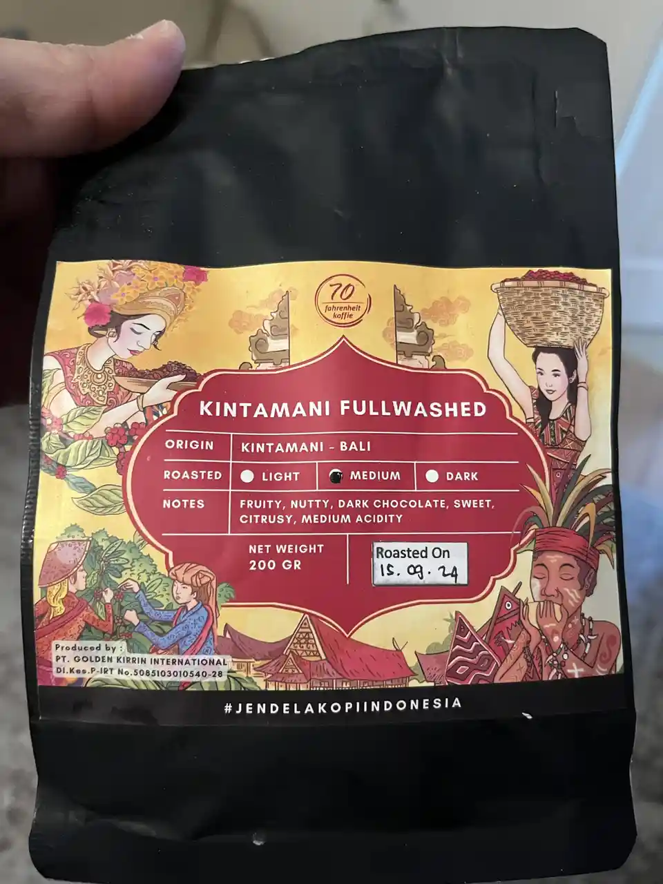 A hand holds a black coffee bag with a colorful label depicting Balinese women in traditional attire harvesting and…