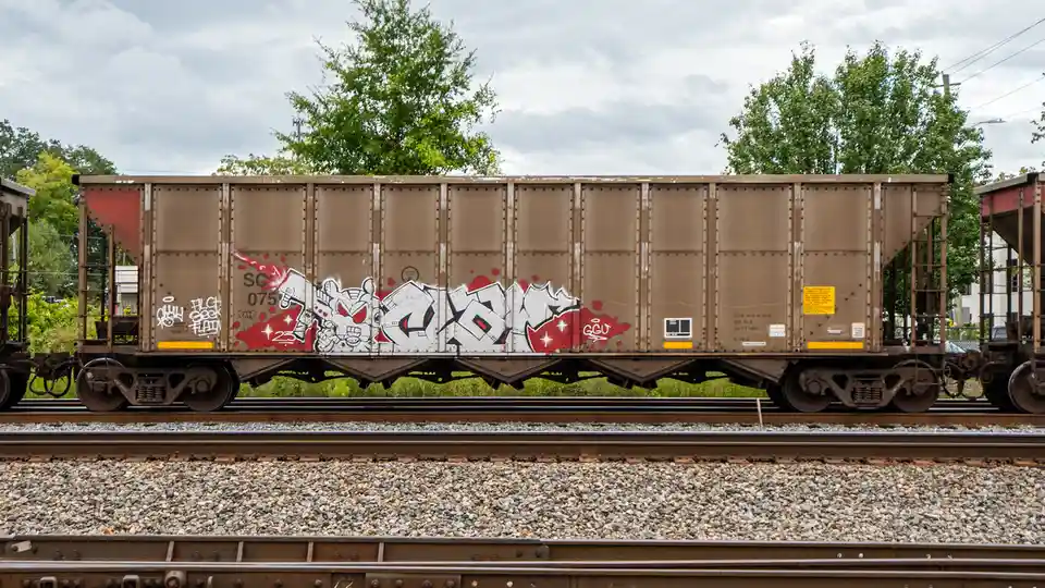 A brown freight train car sits on tracks under a cloudy sky, covered in graffiti including a cartoon character and the text…