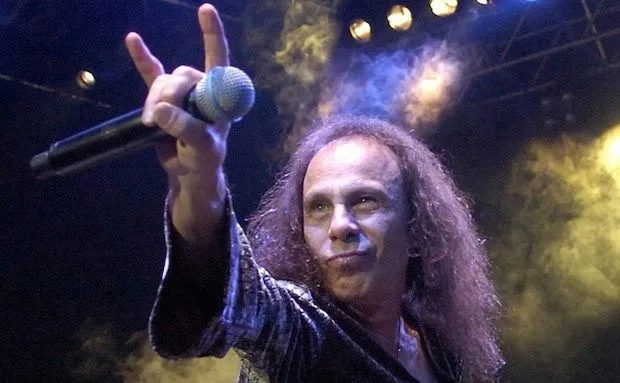 A man with long curly hair and a shiny black jacket holds a microphone in his right hand, making a rock hand gesture with…