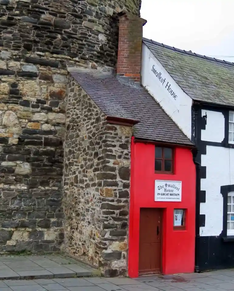 A tiny stone house with a bright red front door and a sign reading 'The Smallest House IN GREAT BRITAIN' is squeezed between…