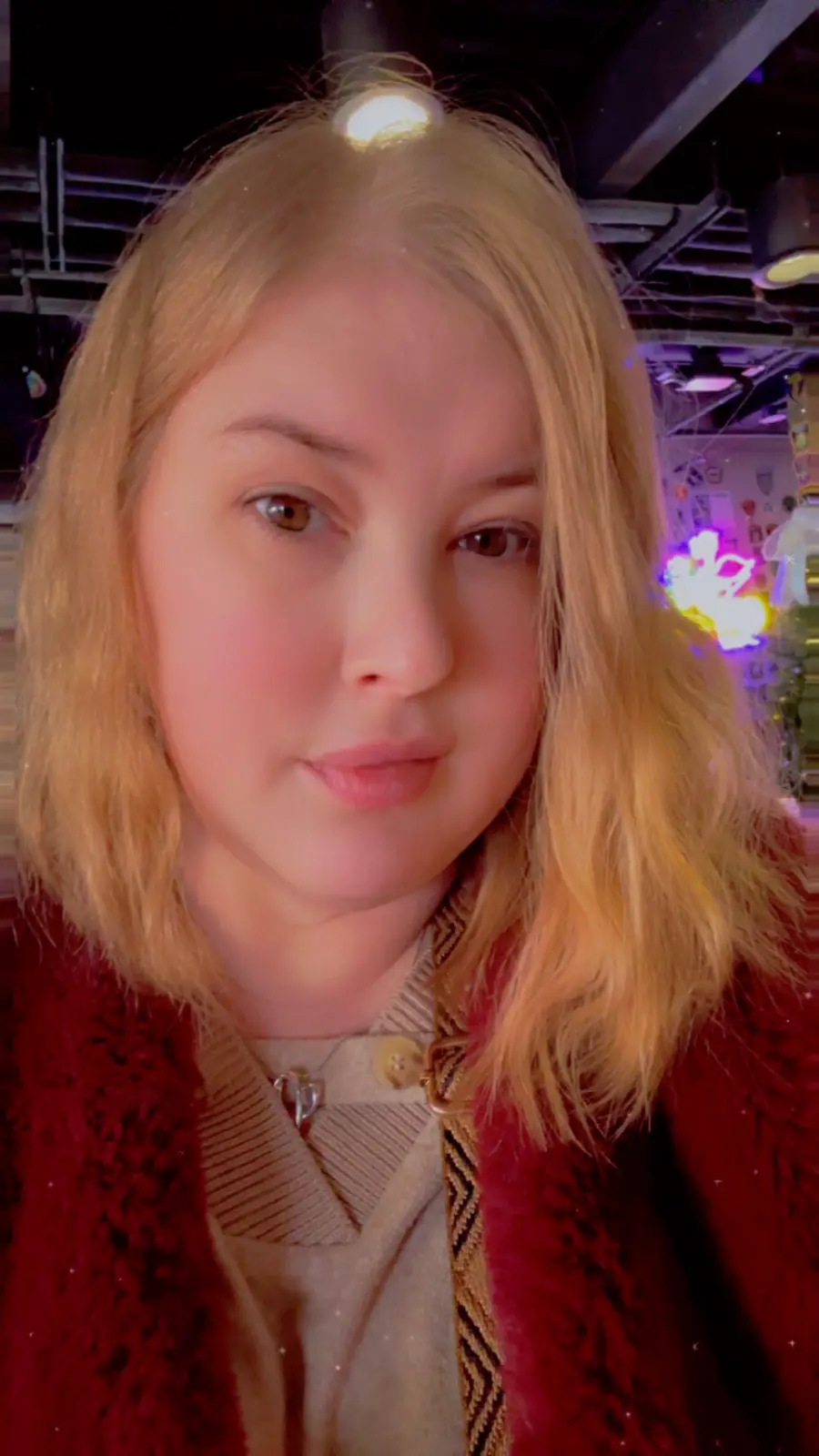 A selfie of a young woman with blonde hair wearing a red fuzzy jacket over a beige top, looking directly at the camera with…
