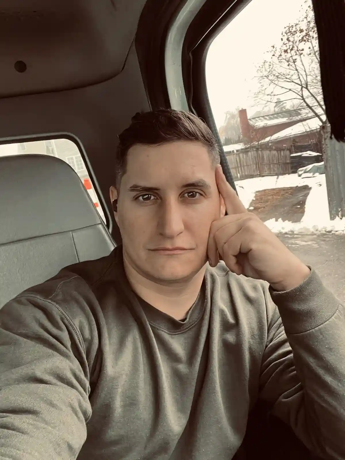 A man sits inside a vehicle, looking directly at the camera with a bored or tired expression, his hand resting against his…