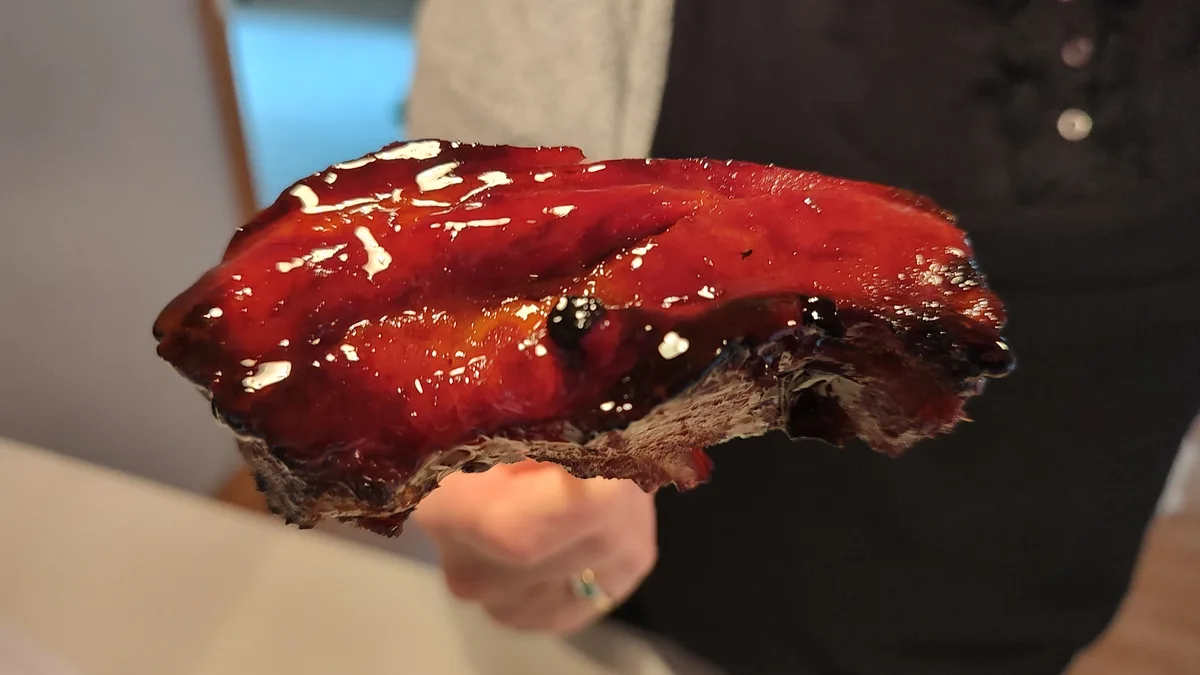 a person holds up a large, glistening piece of meat covered in a thick, dark red sauce, with white specks and charred edges…