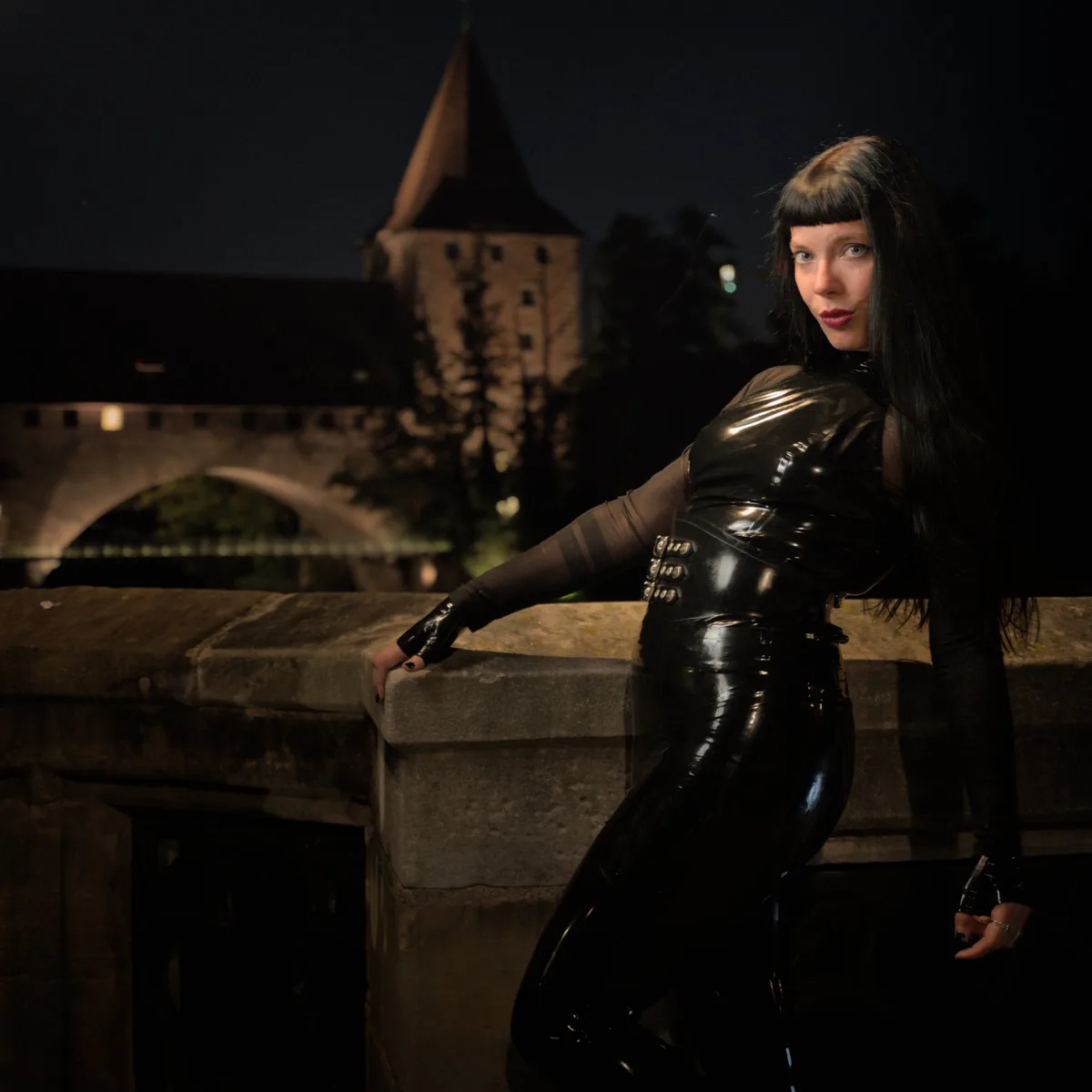 A woman with long black hair and bangs, wearing a shiny black latex catsuit with a corset-style belt, leans against a stone…