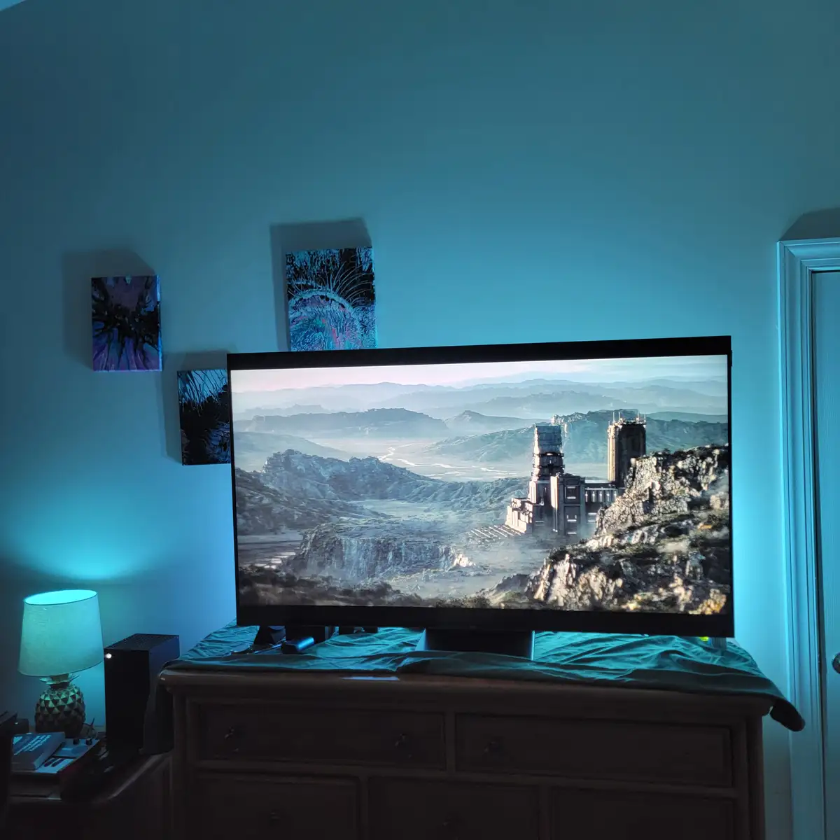 A TV in a dimly lit room displays a vast, desolate fantasy landscape with ancient ruins on a cliffside. The room has blue…