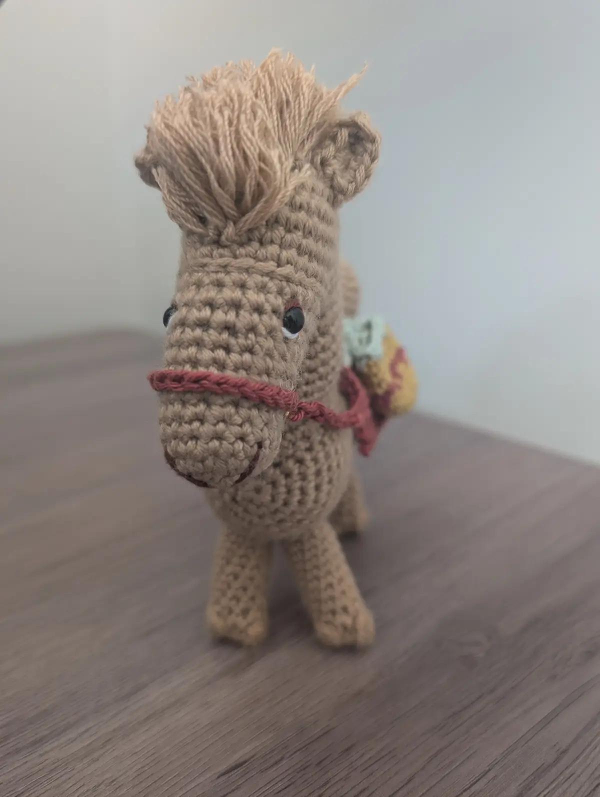 A handmade, beige crocheted horse toy with a messy, spiky mane, black button eyes, and a red yarn bridle stands on a wooden…