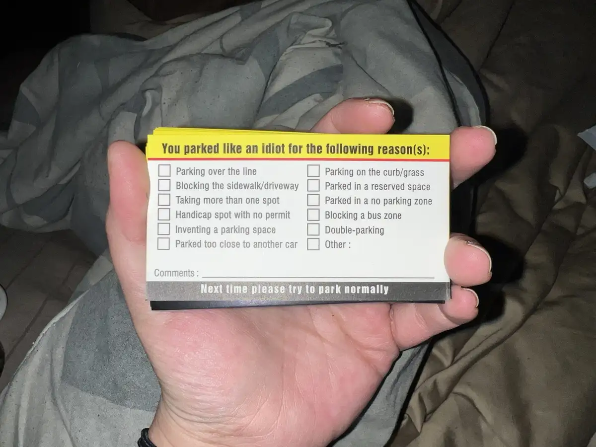 A hand holds a yellow and white parking violation notice card listing common parking infractions like 'parking over the…