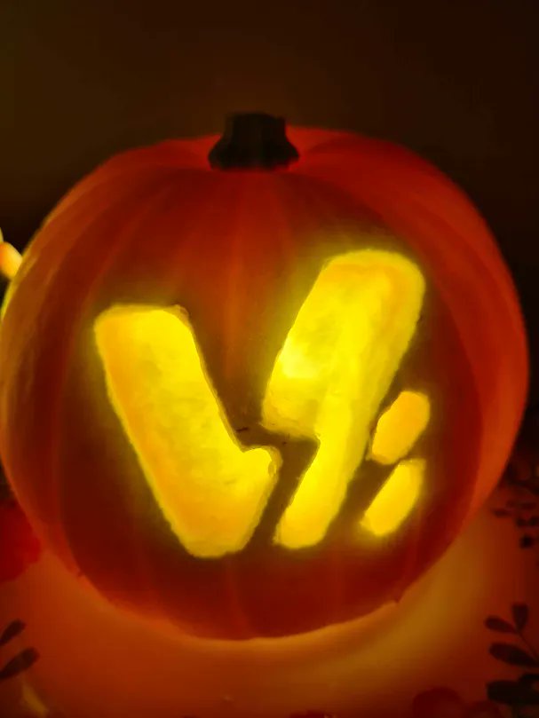 A close-up of a carved Halloween pumpkin with a glowing interior, its face shaped to resemble the letter 'V' followed by a…