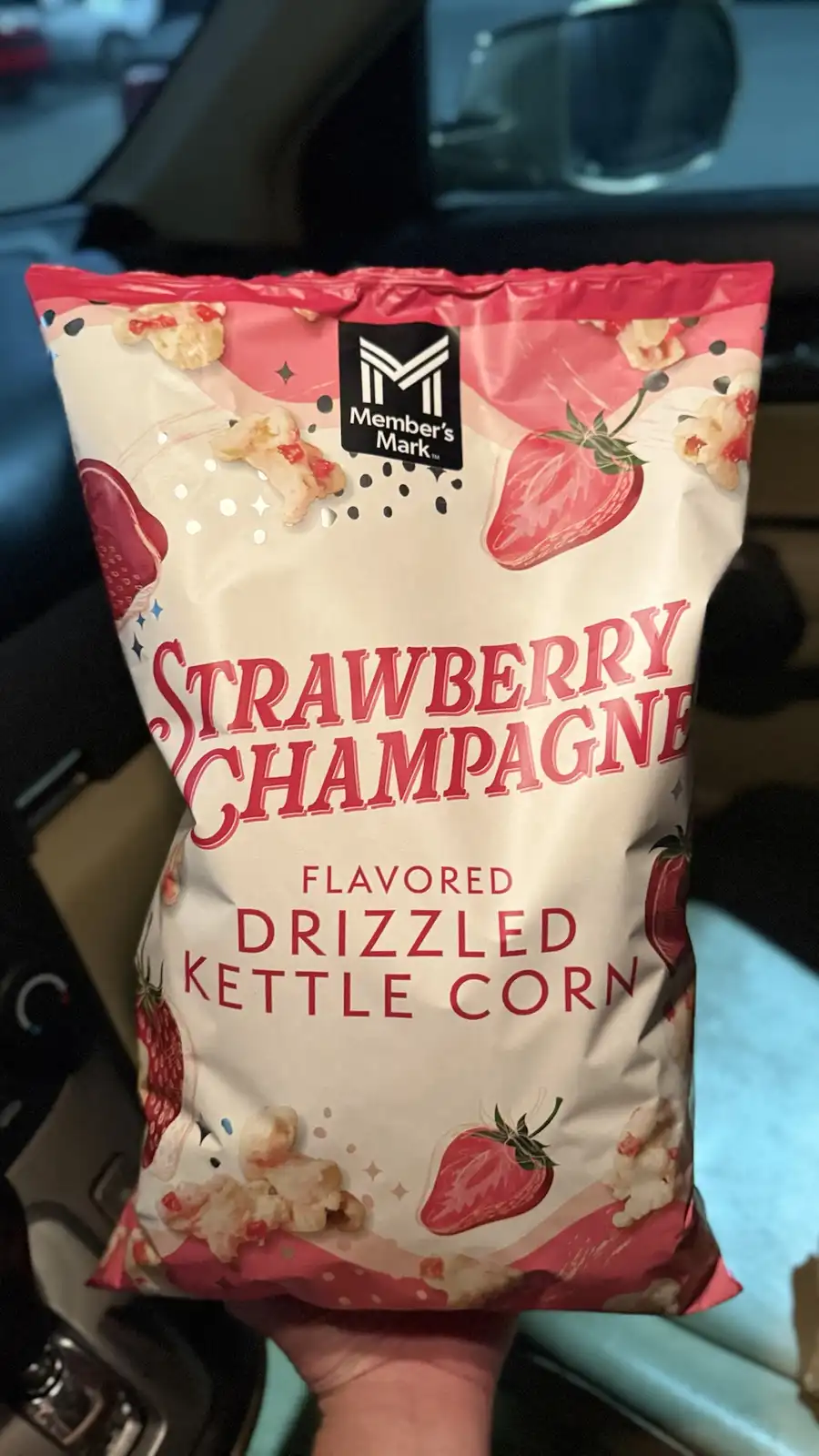A hand holds a bag of Member's Mark Strawberry Champagne Flavored Drizzled Kettle Corn, featuring pink and white packaging…