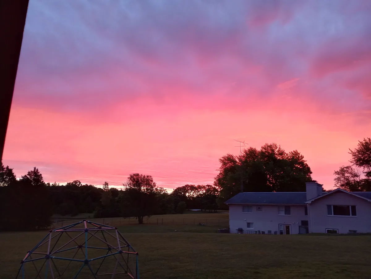 A photo of a vibrant pink and purple sunset over a suburban backyard with a house, trees, and a colorful geodesic dome…