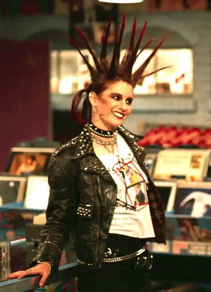 A woman with a tall, spiky red-and-black mohawk hairstyle, wearing a studded black leather jacket over a graphic t-shirt…