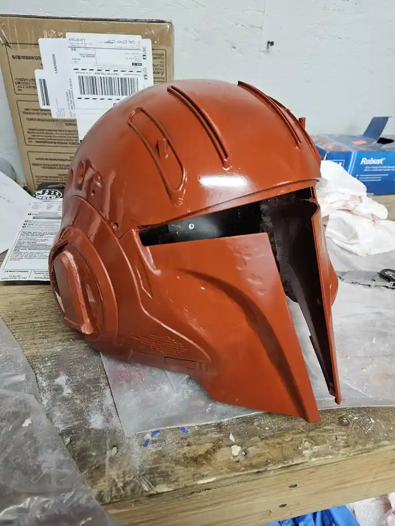 A freshly painted orange helmet, resembling The Mandalorian's, sits on a cluttered workbench surrounded by paint supplies…
