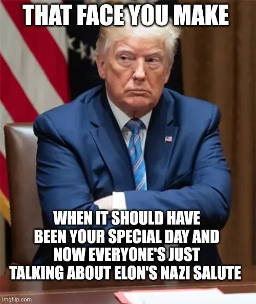 A photo of Donald Trump sitting at a desk with arms crossed, looking displeased. An American flag is visible behind him. The…