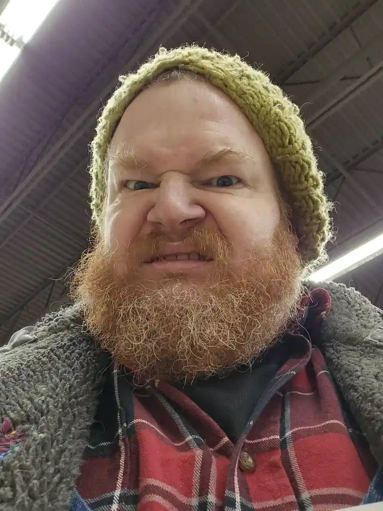 A man with a thick reddish-brown beard and a green knitted beanie makes a scrunched, unimpressed face at the camera. He…