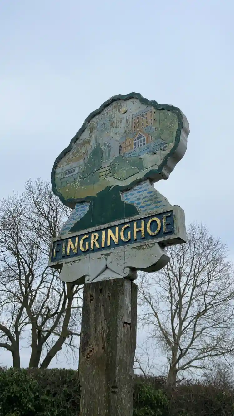 A weathered, hand-painted village sign for 'Fingringhoe' features a faded illustration of local buildings and landscape…
