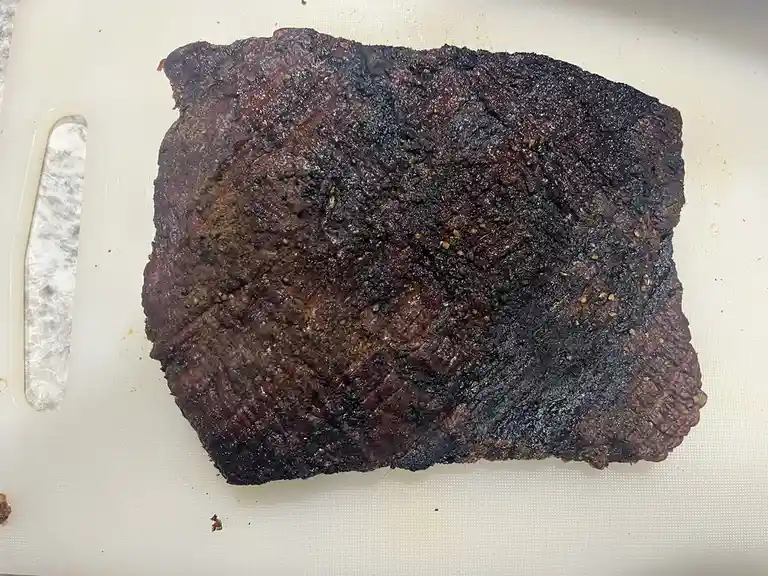 A large, heavily charred piece of meat rests on a white cutting board, its surface blackened and unevenly cooked, suggesting…