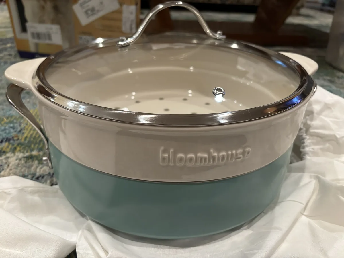 A two-toned ceramic pot with a glass lid and metal handles sits on a white cloth, branded with the word 'bloomhouse'…