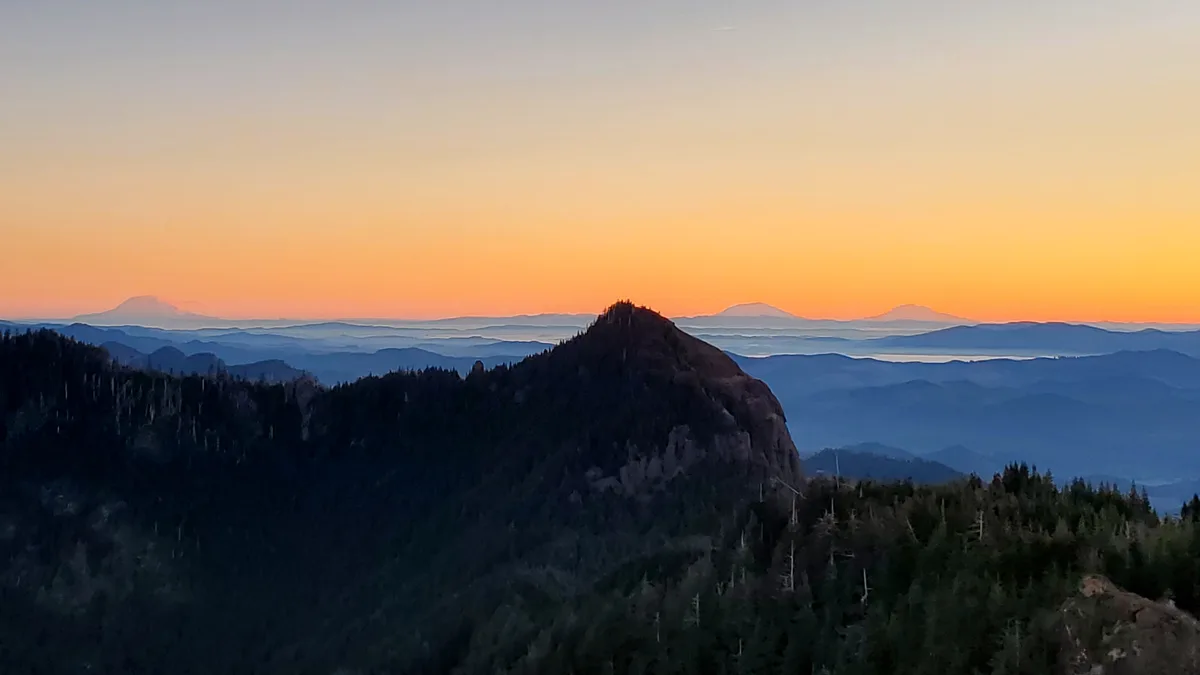 A scenic mountain landscape at sunrise or sunset, featuring layered ridges and distant peaks under an orange and blue sky…