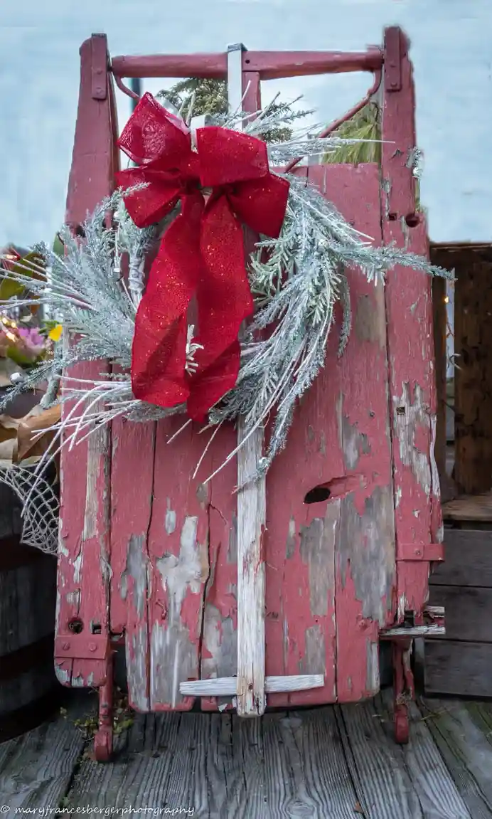 A weathered, red-painted wooden sled with peeling paint stands upright on a wooden deck, adorned with a large red glittery…