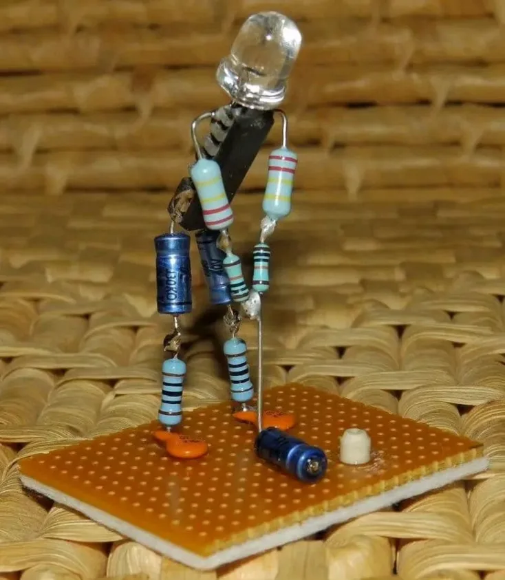 A tiny humanoid figure constructed from electronic components like resistors, capacitors, and a LED for a head, standing…