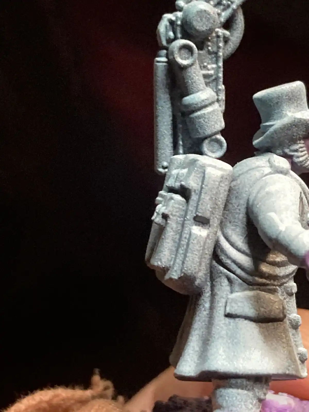 A close-up of a grey-painted miniature figurine of a man in a top hat and coat, heavily laden with complex steampunk-style…