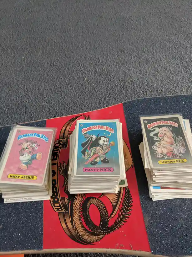 Three stacks of vintage Garbage Pail Kids trading cards are arranged on a skateboard deck featuring a red and black dragon…