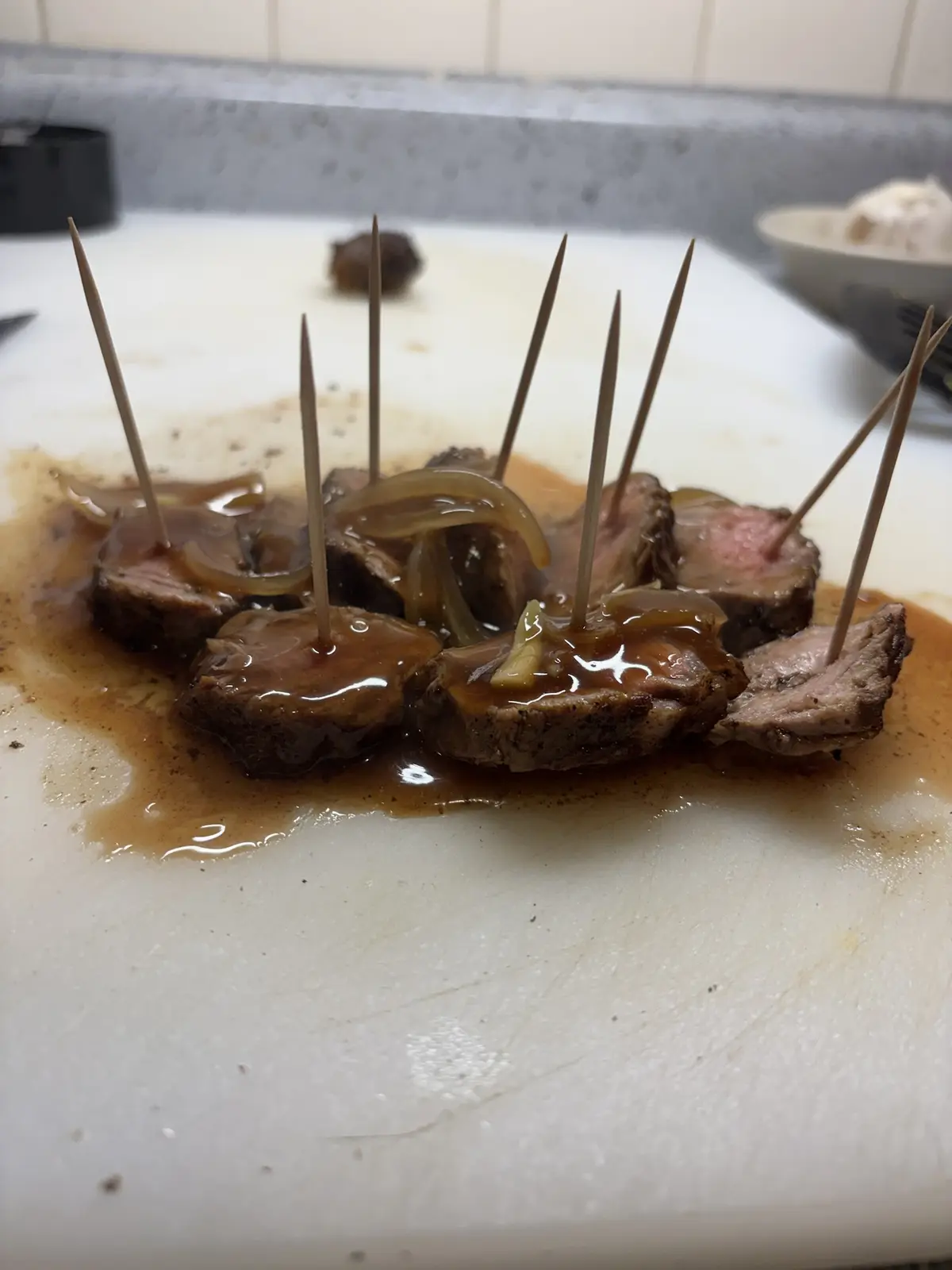 Sliced cooked steak pieces on a white cutting board, each pierced with multiple wooden toothpicks, covered in sauce and…