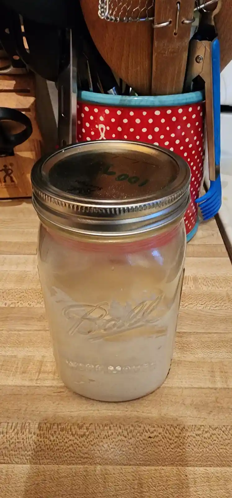 A close-up photo shows a glass Ball mason jar with a metal lid, sitting on a wooden countertop. The lid has the word 'Look'…
