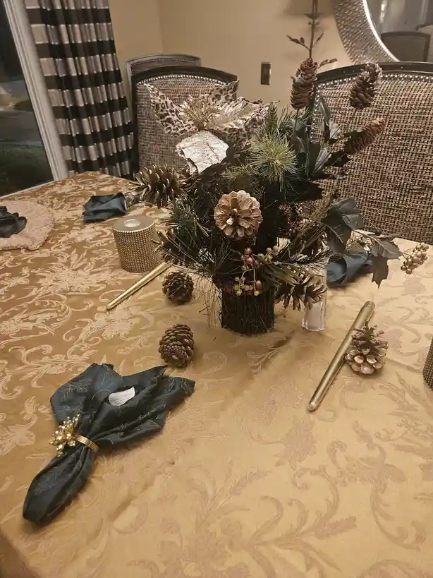 A dining table is set with a gold patterned tablecloth, dark blue napkins with gold napkin rings, and a centerpiece made of…
