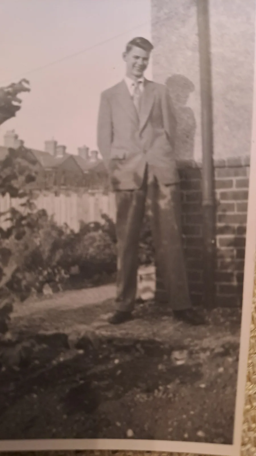 A grainy, sepia-toned vintage photograph shows a young man in a suit and tie standing outdoors next to a brick wall…