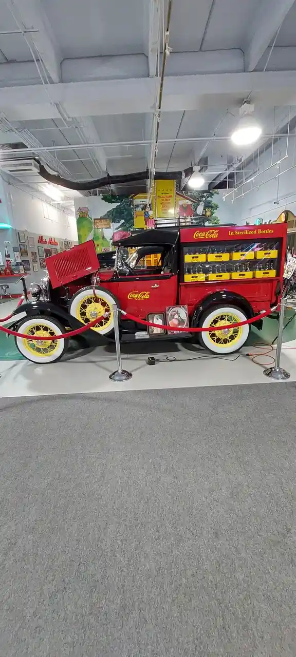A vintage red Coca-Cola delivery truck from the 1920s or 1930s is displayed in a museum, featuring yellow wheels, a black…