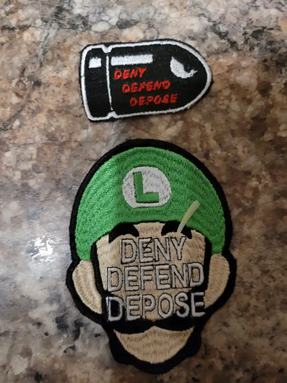 Two embroidered patches lie on a speckled countertop: one shaped like a bullet with red text 'DENY DEFEND DEPOSE', and…