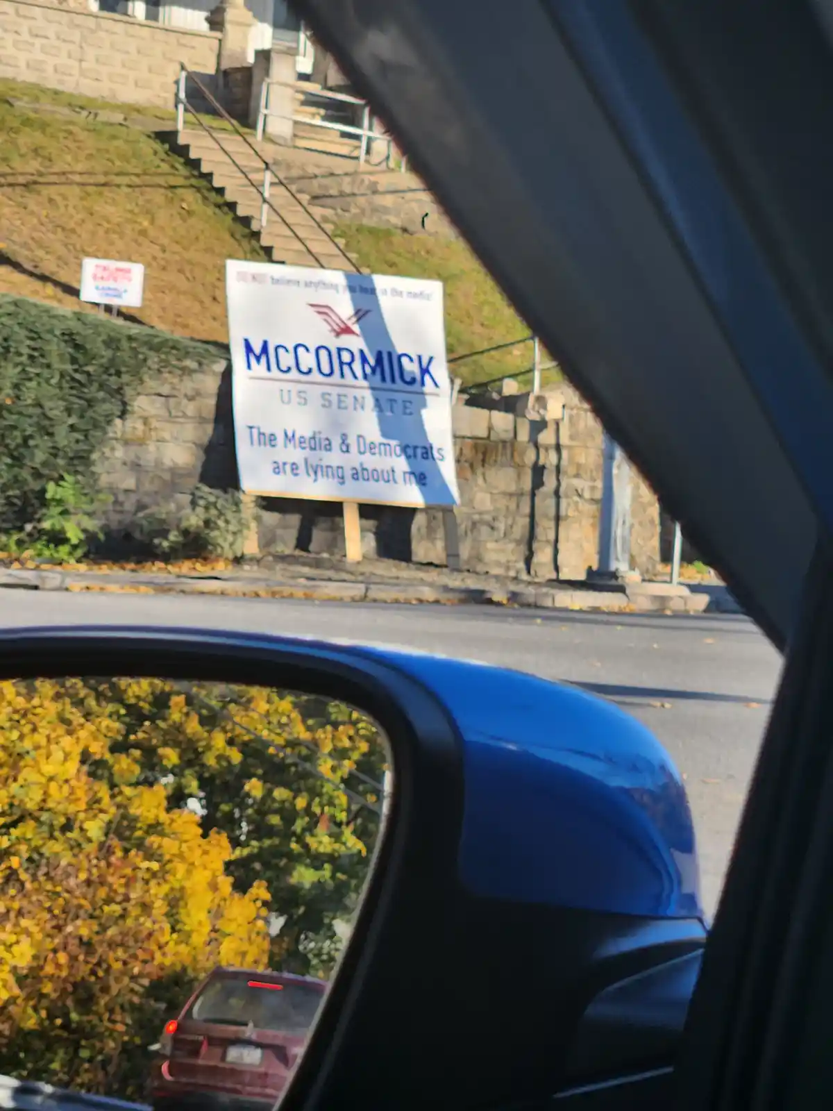 A photo taken from inside a car shows a political campaign sign for 'McCormick US Senate' standing on a grassy slope next to…