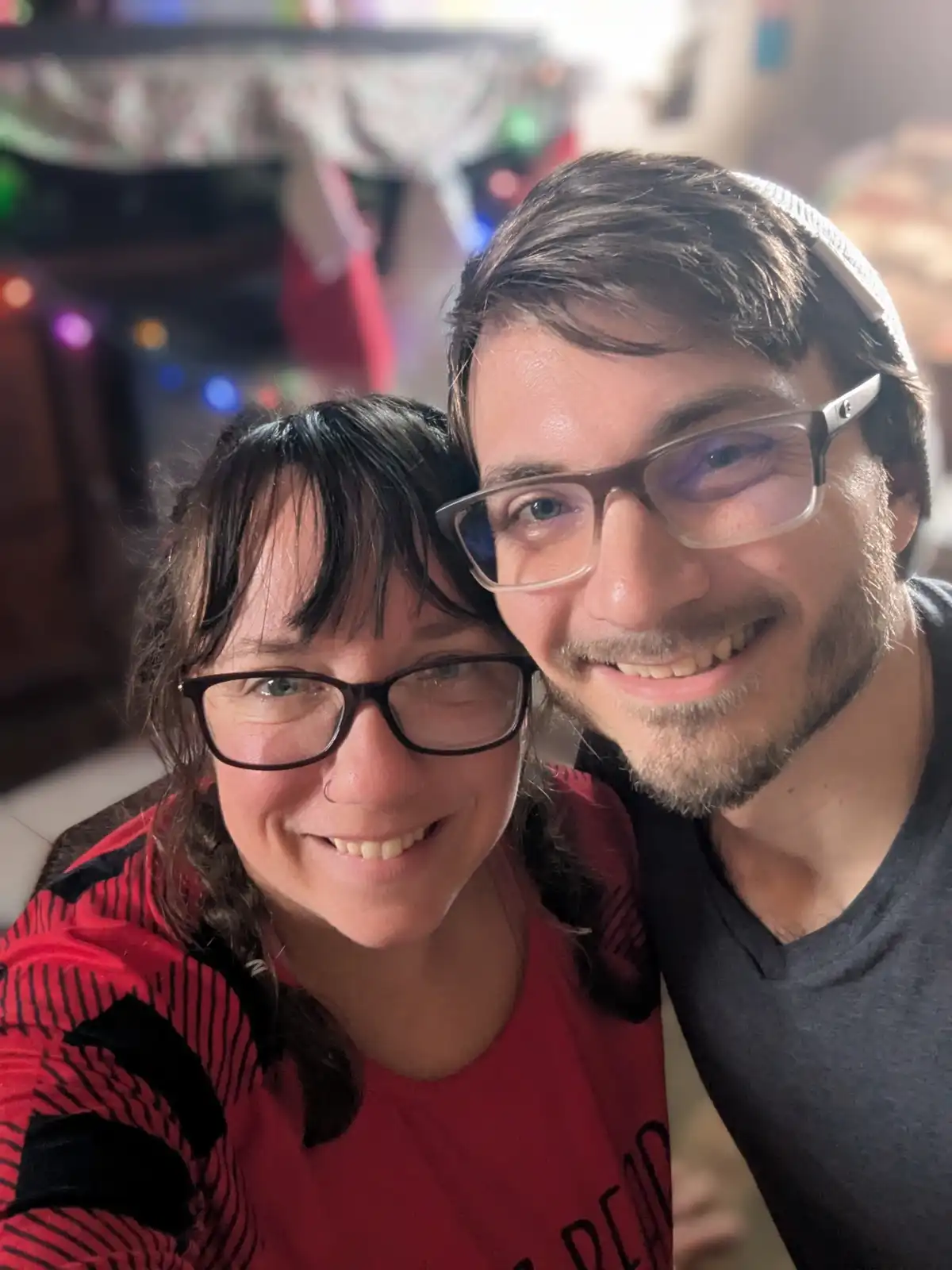 A selfie of a smiling man and woman with glasses, the man has his arm around the woman. The background is blurry with…