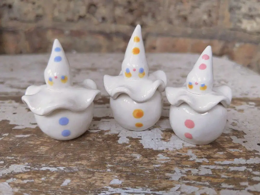 Three small white ceramic figurines shaped like witches with pointed hats and wavy hoods sit on a weathered wooden surface…