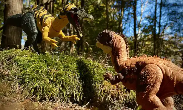 Two plastic dinosaur toys, one yellow and black striped and one brown, are posed facing each other on a mossy forest floor…
